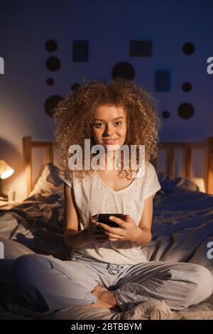 Portrait vertical complet de la jeune femme aux cheveux bouclés assise sur le lit tôt le matin et souriant à l'appareil photo avec joie Banque D'Images