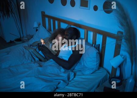 Portrait grand angle de jeunes couples de race mixte, allongé au lit la nuit, regardant l'écran du smartphone tout en parlant, espace de copie Banque D'Images