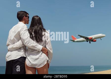 petite amie en bikini et petit ami en chemise blanche se tenir près de l'immense océan calme et regarder l'avion sous le ciel bleu vue arrière Banque D'Images