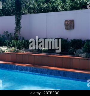 Tête de Lion fontaine avec l'eau s'écoulant dans une petite piscine dans une tendance bien soulevée frontière avec des arbustes bas à côté d'une piscine Banque D'Images