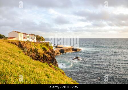 Falaises au bord de l'océan Atlantique dans le village de pêcheurs Lagoa, île de Sao Miguel, Açores, Portugal. Maisons traditionnelles sur la colline au-dessus de la mer. Lumière du coucher du soleil. Photo horizontale. Banque D'Images