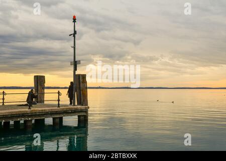 Vue panoramique sur le lac avec un couple prenant des photos sur une jetée en bois au coucher du soleil, Lac de Garde, Lazise, Vérone, Vénétie, Italie Banque D'Images
