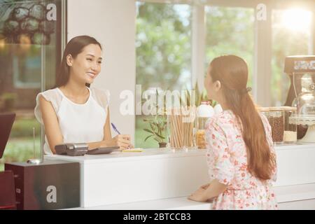 Belle femme souriante Barista debout au comptoir et écrivant l'ordre des jeunes clients Banque D'Images