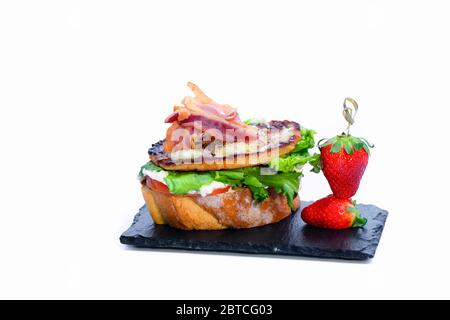 Gros plan sur un délicieux sandwich ouvert avec assortiment de viande, légumes et fromage à côté des fraises fraîches sur une assiette sombre sur fond clair. Spani Banque D'Images