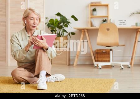 Femme sérieuse et mûre assise sur le sol et travaillant en ligne sur une tablette numérique à la maison Banque D'Images