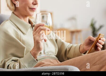 Gros plan d'une femme mûre assise sur un fauteuil avec un verre de vin blanc et utilisant son téléphone portable Banque D'Images