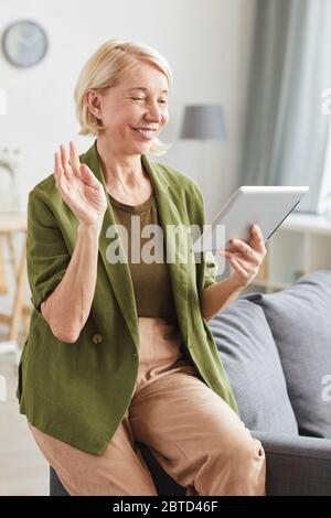 Femme âgée vêtue de vêtements décontractés regardant une tablette numérique et souriant à son ami lorsqu'elle est assise à la maison Banque D'Images