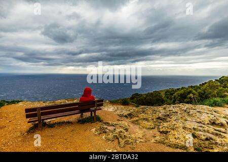 Banc sur la falaise de mer sur Malte avec une veste rouge à capuchon femme sur un jour de tempête nuageux Banque D'Images