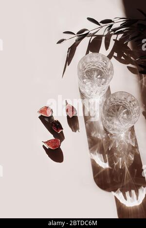 Scène de la vie d'été. Deux verres d'eau, cocktails avec de longues ombres, figues coupées fruits et feuilles d'oliviers sur fond beige table en plein soleil Banque D'Images