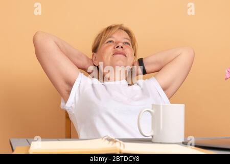 Femme d'âge moyen en t-shirt blanc assis à la table, tient ses mains sous la tête et montre des aisselles coiffées non rasées Banque D'Images