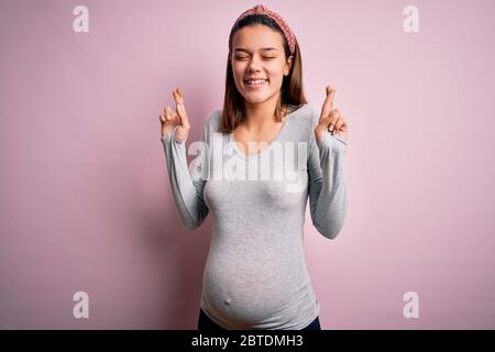 Jeune belle adolescente fille enceinte s'attendant à bébé sur fond rose isolé gesting doigt traversé sourire avec espoir et les yeux fermés. Chance a Banque D'Images