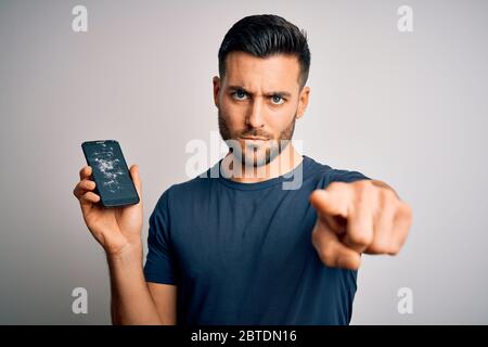 Jeune homme beau tenant un écran de smartphone cassé et fissuré sur un arrière-plan isolé pointant avec le doigt vers l'appareil photo et vers vous, signe de main, pos Banque D'Images