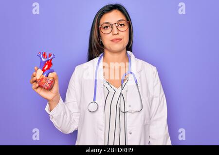 Jeune médecin spécialiste en cardiologie, femme tenant le cœur médical sur fond de pruple avec une expression confiante sur le visage intelligent pensant sérieux Banque D'Images