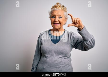 Senior belle femme à cheveux gris portant couronne de reine d'or sur fond blanc souriant et confiant gestant avec la main faisant de petites dimensions signe wi Banque D'Images