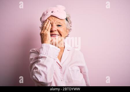 Femme âgée belle portant un masque de sommeil et un pyjama sur fond rose isolé couvrant un œil avec la main, sourire confiant sur le visage et em surprise Banque D'Images