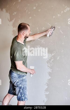 Mur de plâtrage mâle avec couteau à mastiquer, gros plan. Fixation de la surface murale et préparation de la peinture. Jeune homme avec une barbe dans un T-shirt et un short enduit de peinture Banque D'Images