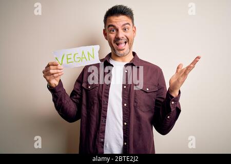 Jeune homme de veggie beau tenant le papier avec le message vegan sur fond blanc très heureux et excité, l'expression du gagnant célébrant la victoire screami Banque D'Images