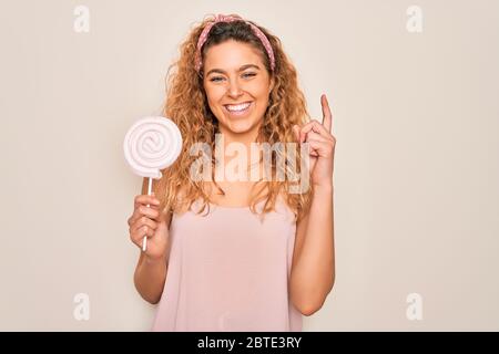Jeune belle femme blonde avec les yeux bleus mangeant sucrée lolipop sucrerie sur fond rose surpris par une idée ou une question pointant du doigt avec h Banque D'Images