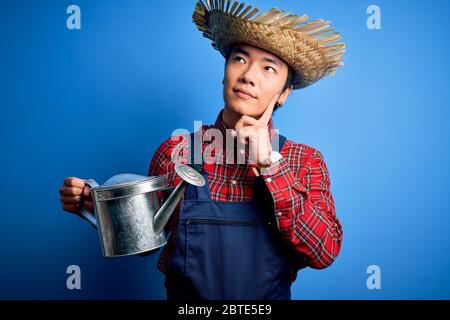 Un jeune fermier chinois charmant portant un tablier et un chapeau de paille portant un arrosoir peut sérieusement faire face à la question, idée très confuse Banque D'Images