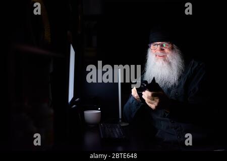 Joyeux homme hippster mûr en utilisant le téléphone tout en travaillant des heures supplémentaires à la maison dans l'obscurité Banque D'Images