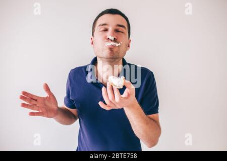 Jeune homme isolé sur fond blanc. Manger délicieux délicieux gras petit gâteau. Une partie de la crème est sur son nez et son visage. Plaisir délicieux Banque D'Images