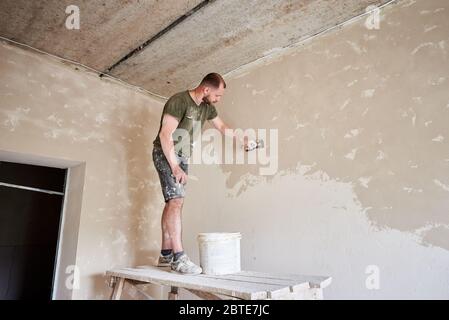 Le constructeur masculin est debout sur un support en bois et travaille avec une spatule avec du plâtre sur le mur dans une petite pièce. Des murs en poutlage à l'intérieur. Un travailleur avec une barbe dans un t-shirt et un Jean est enduit de peinture Banque D'Images