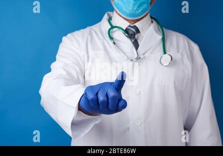 un médecin adulte de sexe masculin, portant un manteau médical blanc et des gants en latex bleu, se tient sur un fond bleu et, avec sa main droite, étire son index vers l'avant Banque D'Images