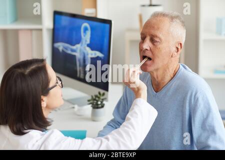 Vue en grand angle chez la femme médecin vérifiant la gorge du patient âgé pendant son examen pendant la consultation en clinique Banque D'Images