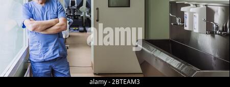 Bras croisés sur la poitrine d'un médecin debout à la fenêtre de la salle de désinfection. Un grand évier en acier inoxydable pour se laver les mains avant la chirurgie. Banque D'Images
