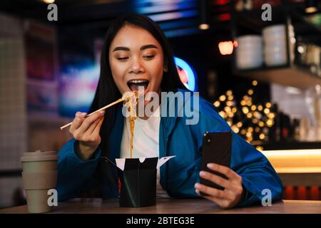 Photo de joyeuse femme asiatique de manger des nouilles chinoises et d'utiliser un téléphone portable tout en étant assise dans un café Banque D'Images