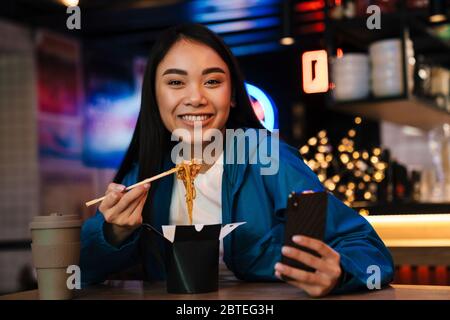 Photo de joyeuse femme asiatique de manger des nouilles chinoises et d'utiliser un téléphone portable tout en étant assise dans un café Banque D'Images