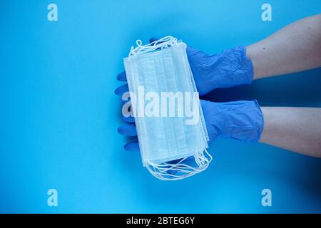 Deux mains d'un médecin en gants médicaux tient, prend, donne des masques chirurgicaux médicaux pour couvrir la bouche et le nez. Vue du dessus, gros plan, sur fond bleu Banque D'Images