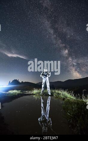 Une longueur complète d'astronaute en costume d'espace jouant de la guitare sous le ciel nocturne avec Milky Way. Guitariste cosmonaute avec instrument de musique réfléchissant dans un petit lac tout en se tenant dans une vallée herbeuse illuminée. Banque D'Images