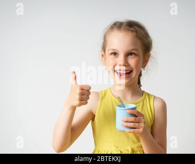 Bonne petite fille blonde en vêtements jaunes est ravie avec la nourriture savoureuse montrant sourire brillant et le pouce vers le haut geste. Portrait de gros plan isolé sur blanc Banque D'Images