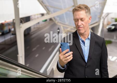 Homme d'affaires mature utilisant le téléphone en allant vers l'escalier roulant dans la ville Banque D'Images