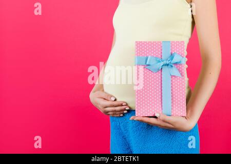 Gros plan de la boîte cadeau rose à pois dans la main de la femme enceinte portant des vêtements colorés à la maison sur fond rose. En attente d'une petite fille. Copier l'espace. Banque D'Images