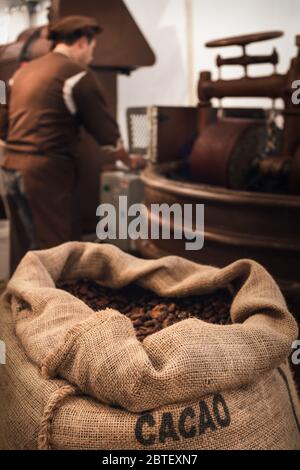 Sac de jute plein de fèves de cacao dans un atelier de chocolaterie, avec un chocolatier mâle travaillant sur l'équipement de conchage et de mélange sur le fond Banque D'Images