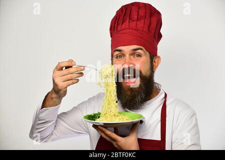 Cuire avec le visage heureux dans l'uniforme bordeaux contient la fourchette et l'assiette. Le chef mange des nouilles italiennes ou asiatiques. Concept de cuisine de restaurant. L'homme avec la barbe tient un plat savoureux sur fond blanc. Banque D'Images