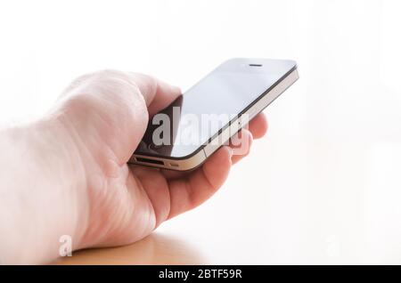 Gros plan de la main d'un homme tenant et touchant ou utilisant le smartphone iPhone 4s sur un bureau en bois sur un arrière-plan blanc lumineux avec espace de copie Banque D'Images