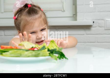 une petite fille repousse émotionnellement une assiette de légumes offerts. les enfants refusent les légumes en faveur de la nourriture de mauvaise qualité Banque D'Images