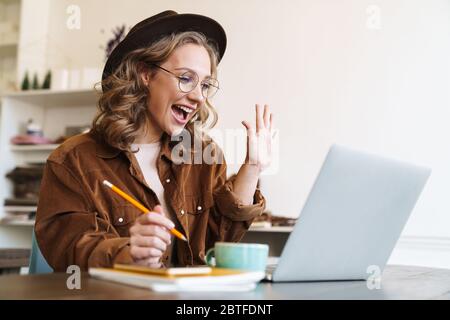 Image d'une jeune femme excitée au chapeau travaillant avec un ordinateur portable et se faisant main tout en étant assise à une table Banque D'Images