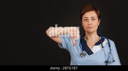 Mécontentement la médecin européenne montre un signe de désapprobation, garde le pouce vers le bas, exprime le mécontentement, fronces de sourcils face au mécontentement, isolé sur fond noir. Concept de langage corporel. Banque D'Images