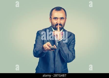 Adulte adulte adulte adulte adulte adulte en costume élégant avec noeud papillon rouge tenant la télécommande de tv et le doigt sur la bouche sur le geste de la ruche. Modèle barbu de race mixte isolé sur Banque D'Images