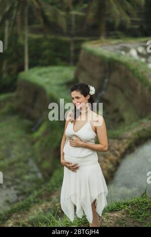 Jeune femme enceinte en robe blanche avec vue sur les rizières en terrasses de Bali dans la lumière du soleil du matin. Harmonie avec la nature. Concept de la grossesse. Banque D'Images