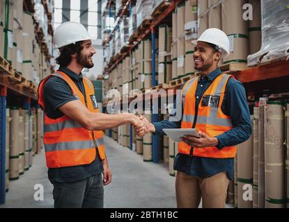 Deux hommes joyeux qui font une poignée de main ferme dans le bureau de logistique tenant une tablette numérique dans un casque de sécurité et un gilet Banque D'Images