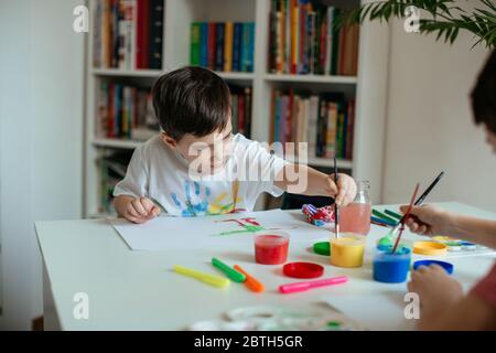 Petit garçon gaucher, qui atteint la peinture avec un pinceau dans la main. Enfant avec pinceau et peintures colorées à la maison. Banque D'Images