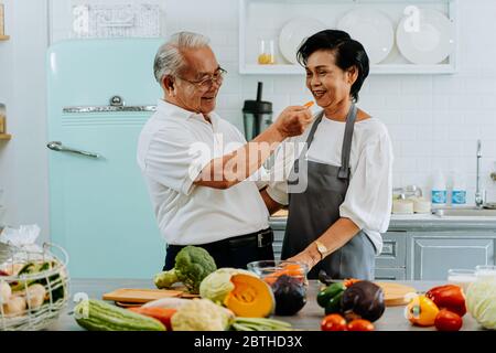 Senior Asian couple de grands-parents ensemble pendant que la cuisson des aliments est l'alimentation de la femme à l'homme à la cuisine. Relation durable concept Banque D'Images