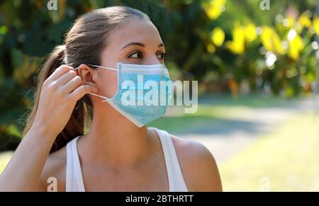 Fille dans le parc met sur un masque médical. Jeune femme sportive portant un masque chirurgical dans le parc de la ville. Copier l'espace. Banque D'Images