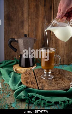 Versez le lait dans le café. Faire du café avec de la crème. Tête en verre à la main. Un verre de cappuccino. Machine à café Geyser sur une table en bois. Faites cuire le petit déjeuner à Banque D'Images