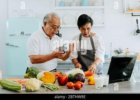 Un couple asiatique senior utilisant un ordinateur portable tout en cuisinant dans la cuisine à la maison. 70 ans homme et femme âgés dans la relation retraite concept de style de vie Banque D'Images
